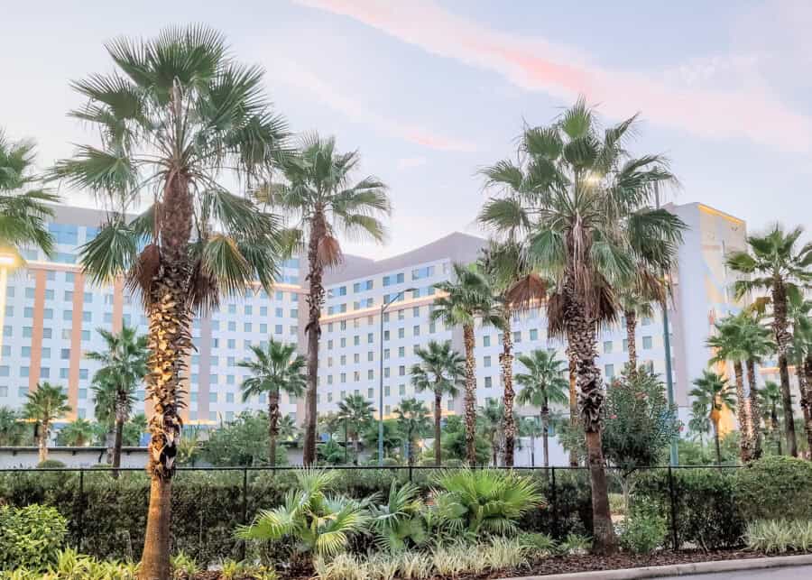 Tall palm trees line a landscaped walkway in front of a large modern hotel building under a soft pastel sunset sky.