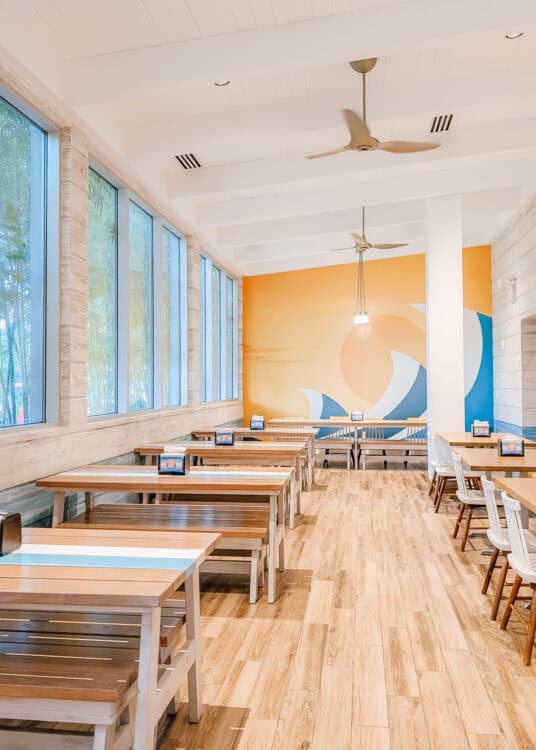 right dining area with long wooden tables and bench seating, large windows letting in natural light, and ceiling fans above. A colorful mural with a stylized wave and sunset decorates the back wall, highlighting the relaxed surf inspired atmosphere.