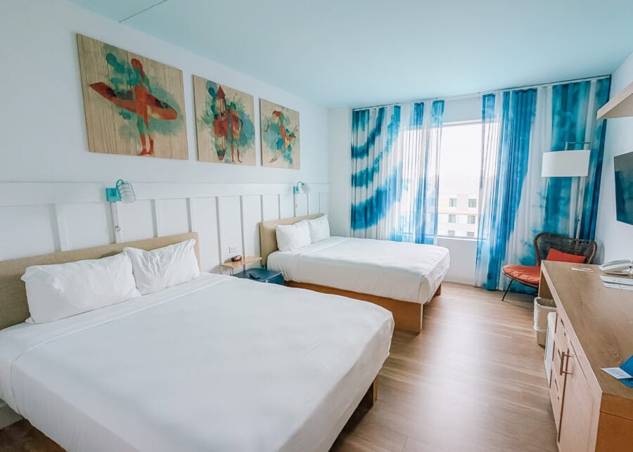 Bright hotel room with two neatly made beds, light wood floors, and blue wave patterned curtains framing a window with an outside view. Surf themed artwork hangs above the beds, reflecting the clean beachy design highlighted in this endless summer surfside review.