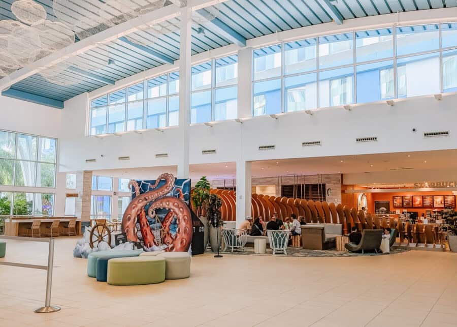 Bright hotel lobby with high ceilings and large windows letting in natural light, featuring a colorful octopus sculpture surrounded by round seating. Guests sit and relax near a cafรฉ area in the background, adding context to the casual atmosphere described in this endless summer surfside review.