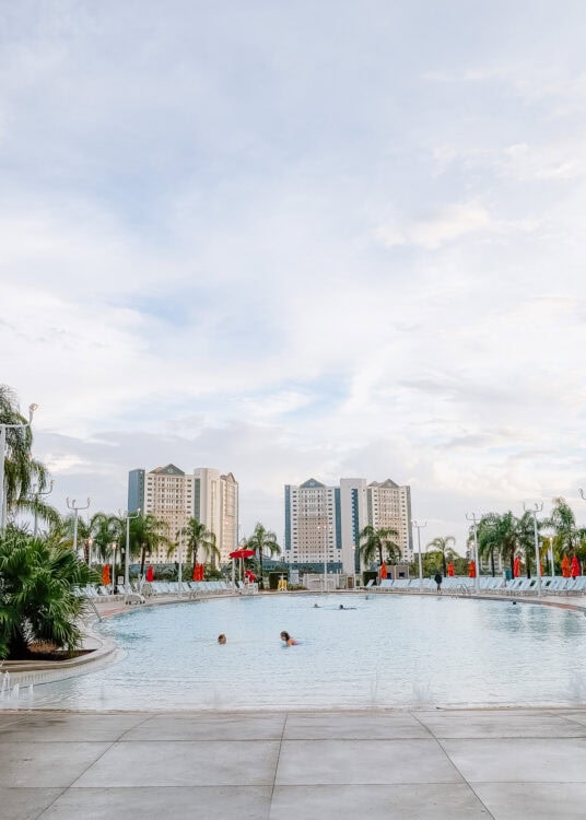 Large outdoor resort pool with guests swimming and relaxing, surrounded by palm trees, lounge chairs, and bright orange umbrellas. Tall hotel buildings rise in the background under a soft cloudy sky, capturing the open airy feel described in this endless summer surfside review.