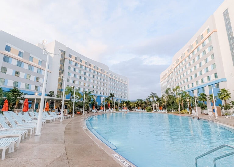 Large resort pool curves between two modern white hotel buildings lined with rows of windows and balconies, with palm trees and lounge chairs surrounding the water. Guests relax poolside under orange umbrellas, capturing the laid back atmosphere highlighted in this endless summer surfside review.