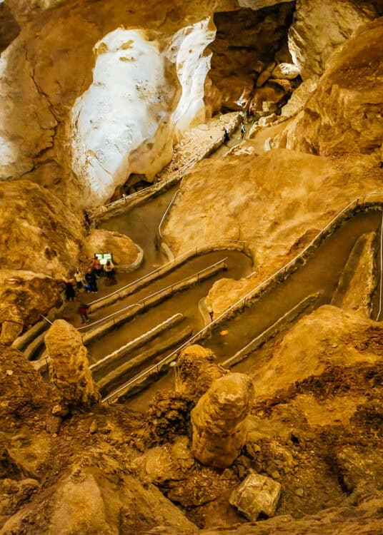 A winding paved trail with metal railings curves through a large cave chamber filled with warm amber lighting and smooth rock formations. Several visitors walk along the switchback path, emphasizing the scale of the cavern walls around them.