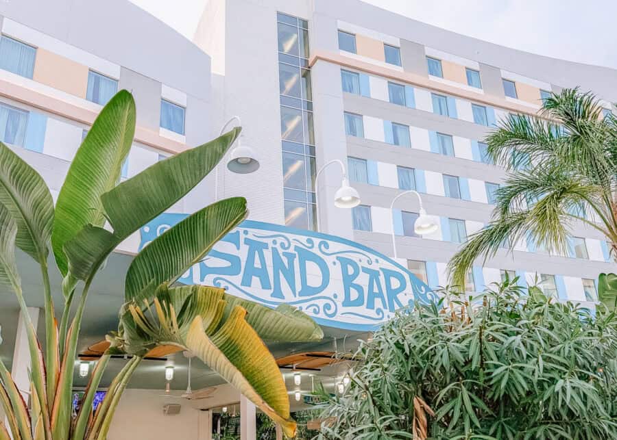 Exterior of a modern hotel building with palm trees and tropical plants in the foreground, featuring a blue sign that reads "Sand Bar" above an outdoor dining area. The beachy decor and casual dining spot reflect the laid back vibe highlighted in this endless summer surfside review.