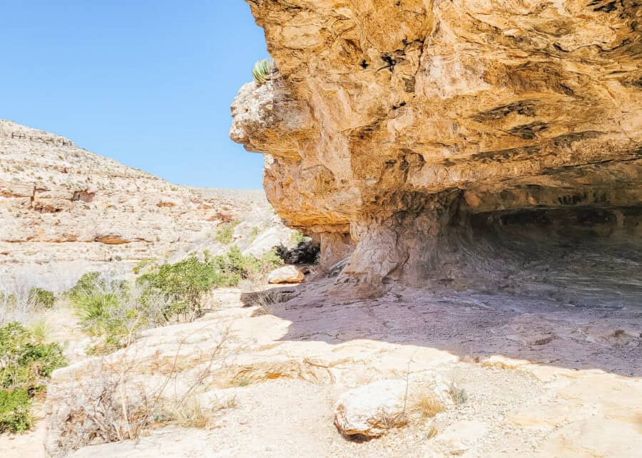 A rocky desert overhang forms a shaded cave along a hiking trail at Carlsbad Caverns, with sunlit cliffs and sparse vegetation stretching into the distance. The rugged landscape and natural shelter add variety to a Carlsbad Caverns itinerary above ground.