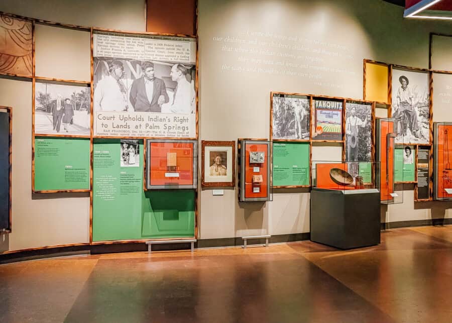 Museum exhibit in Palm Springs featuring historic photographs, newspaper clippings, and cultural artifacts about the Agua Caliente Band of Cahuilla Indians. Large wall displays and framed exhibits highlight Indigenous history and land rights inside the interactive gallery space.