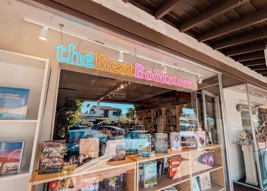 Storefront window of a bookstore with a colorful neon sign reading "the Best Bookstore" above shelves filled with books and displays. Reflections of parked cars and palm trees appear in the glass, blending the cozy interior with the sunny street outside.