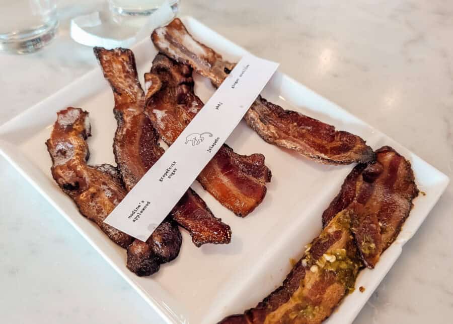 Assorted strips of crispy bacon served on a white rectangular plate with a tasting guide labeling flavors like โNodine's Applewood,โ โGrapefruit Sugar,โ โJalapeรฑo,โ โPB & Jโ and โGinger Scallion.โ One strip is topped with green peppers while the rest have caramelized edges and smoky seasoning.