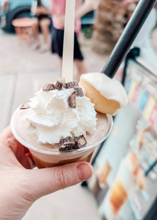 Hand holding a creamy chocolate milkshake topped with whipped cream, chopped chocolate pieces, and a frosted mini doughnut on the rim. The dessert drink is photographed outdoors beside a pastel colored food truck with blurred people and palm trees in the background.