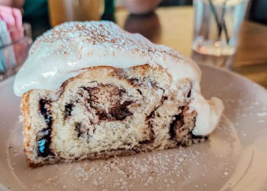 Close up of a thick cinnamon roll with cream cheese icing dripping over the sides and powdered sugar sprinkled on top. The soft pastry is sliced open to show swirls of cinnamon filling and melted chocolate inside.