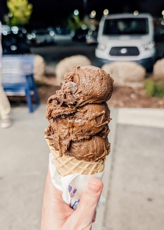 Hand holding a waffle cone stacked with three scoops of rich chocolate ice cream filled with chocolate chunks at night. Blurred lights and parked cars in the background give the dessert shop scene a cozy evening atmosphere.