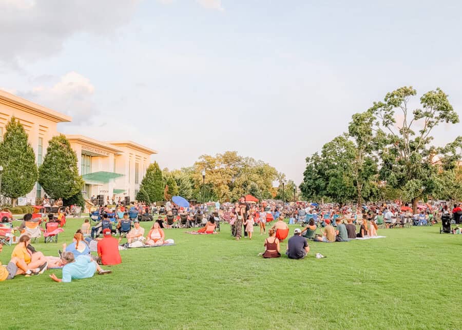 Large crowd gathered on the grassy lawn outside the Huntsville Museum of Art during an outdoor community event in Huntsville, Alabama. Families and groups sit in lawn chairs and blankets under a warm evening sky surrounded by trees