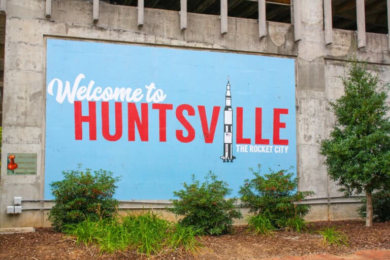Large blue mural reading "Welcome to Huntsville The Rocket City" painted on a concrete wall with a Saturn V rocket illustration between the letters. Small green bushes and landscaping sit in front of the colorful Huntsville sign