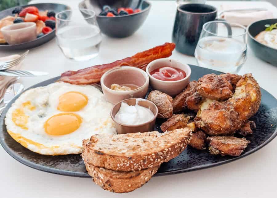 Breakfast plate with two sunny side eggs, crispy bacon, roasted potatoes, and slices of seeded toast arranged on a dark dish. Small bowls of ketchup, butter, and creamy sauce sit alongside the meal with drinks and fruit bowls in the background.