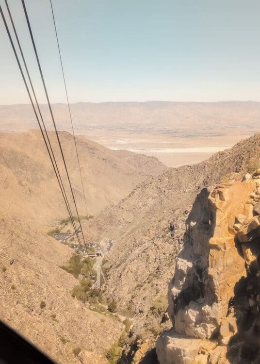 View from a mountain tram looking down over rocky desert cliffs and cable lines stretching into the valley below. Hazy mountains and a wide desert landscape fill the background beneath a pale blue sky.