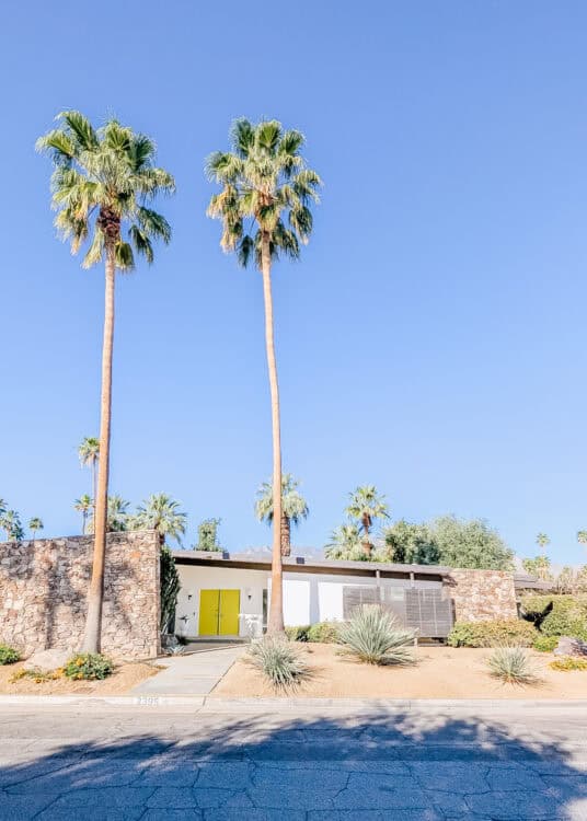 Mid century modern home in Palm Springs with bright yellow front doors, stone walls, and tall palm trees set against a clear blue sky, showcasing iconic architecture among things to do in Palm Springs. Desert landscaping with agave plants and a mountain backdrop adds to the classic California desert aesthetic.