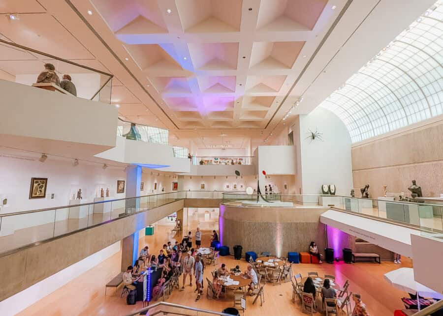 Bright modern museum interior with high ceilings, geometric skylight, and multiple levels of galleries displaying sculptures and artwork. Visitors gather around tables on the main floor while others explore exhibits along the upper walkways.