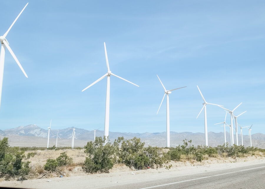 Row of tall white wind turbines standing across a desert landscape with low shrubs and mountains in the distance under a clear blue sky. A road runs along the foreground, emphasizing the wide open and airy setting.