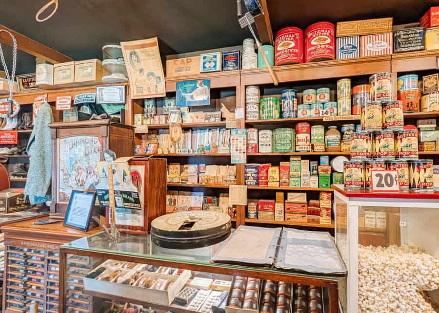 Vintage general store display with wooden shelves filled with colorful tins, boxes, and retro packaging for coffee, popcorn, and pantry goods. A glass counter in the foreground holds small items and an open ledger while a popcorn case labeled "20ยข" adds to the nostalgic atmosphere.