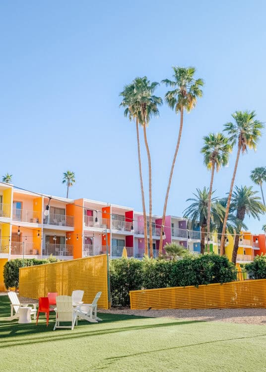 Colorful retro style motel with bright orange, pink, and yellow walls framed by tall palm trees under a clear blue sky. A small grassy courtyard with white lounge chairs and string lights creates a cheerful outdoor gathering space.