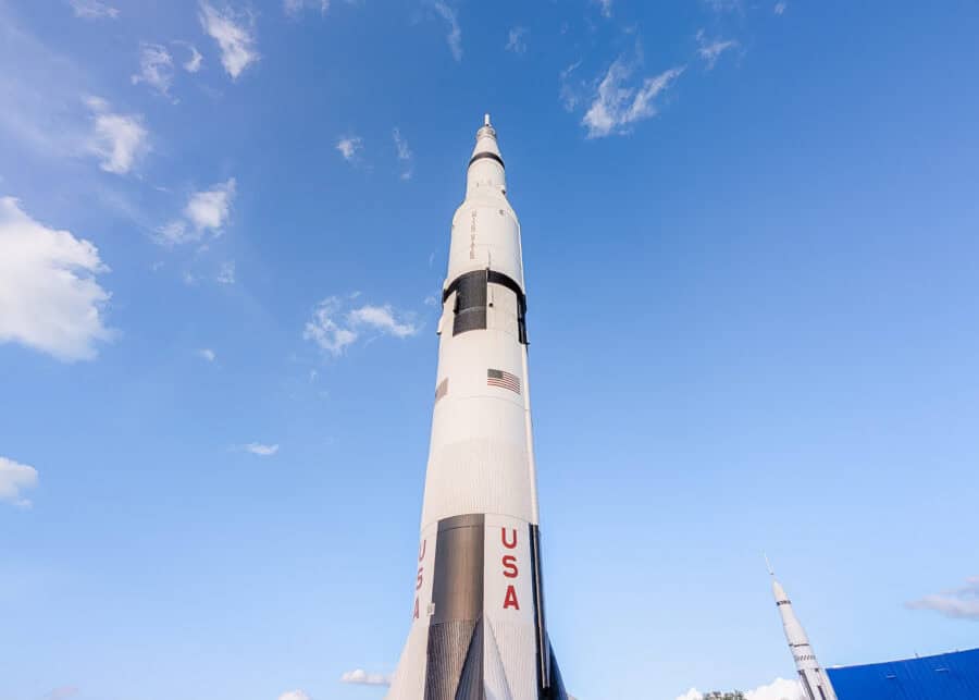 Massive Saturn V rocket standing upright against a bright blue sky at the U.S. Space and Rocket Center in Huntsville. The white rocket with bold red "USA" lettering towers above visitors and nearby exhibits, showcasing the cityโs space exploration history.