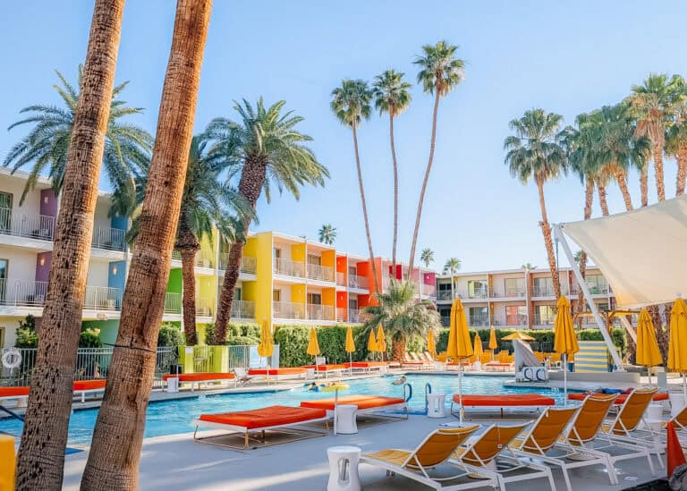 Colorful mid century style hotel in Palm Springs with rainbow painted balconies surrounding a sunny pool lined with yellow umbrellas and lounge chairs, capturing a vibrant scene of things to do in Palm Springs. Tall palm trees tower over guests relaxing by the water, highlighting the laid back desert resort atmosphere.