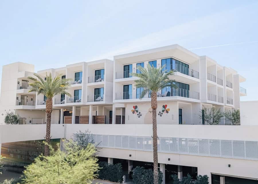 Modern white hotel or apartment building with glass balconies and tall palm trees in Palm Springs under a clear blue sky. The sleek desert style architecture and sunny weather create a relaxed luxury resort atmosphere.