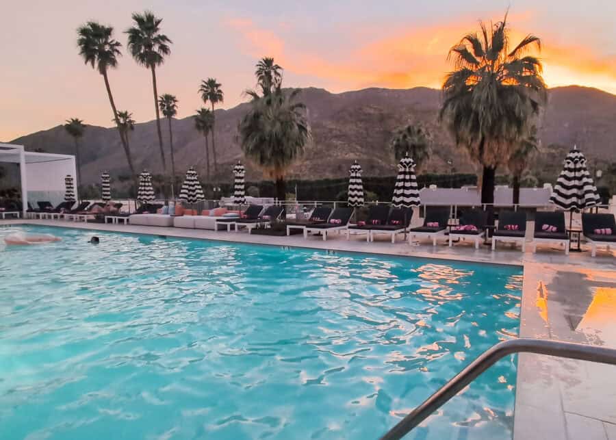 Outdoor pool at sunset with turquoise water reflecting warm orange and pink skies behind a line of palm trees and mountains. Black and white striped umbrellas and lounge chairs surround the pool, creating a relaxed desert resort atmosphere.