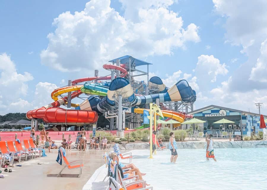 Colorful twisting water slides tower above a crowded wave pool at an outdoor water park on a sunny day. Families relax in lounge chairs and splash in the shallow water beneath bright blue skies filled with fluffy clouds.