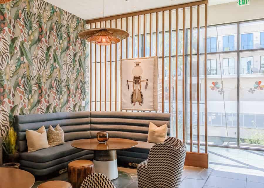 A stylish hotel lounge with curved blue banquette seating, patterned chairs, tropical botanical wallpaper, and a woven pendant light creates a cozy gathering space beside floor-to-ceiling windows. Natural light fills the room while views of the courtyard and modern architecture outside complement the contemporary desert-inspired design.