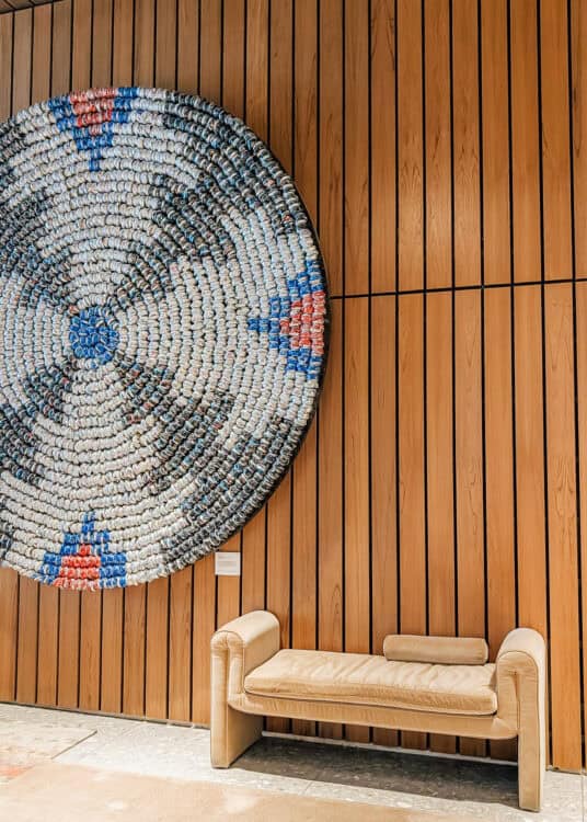 A tan upholstered bench sits beneath a large circular wall sculpture made from tightly arranged recycled bottle caps in white, blue, black, and red tones against a warm wood slat wall. The modern art installation creates a striking focal point in the hotel lobby with clean lines and minimalist seating below it.