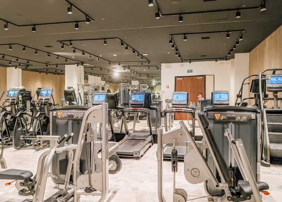 A modern hotel fitness center with rows of treadmills, strength training machines, and cardio equipment arranged in front of mirrored walls under black track lighting. The spacious gym offers a clean, well-equipped workout space with multiple exercise stations and large reflective surfaces that make the room feel even larger.
