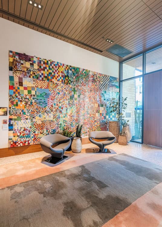 Contemporary lobby at Thompson Palm Springs featuring two sculptural lounge chairs placed in front of a large colorful mosaic wall installation. Floor to ceiling windows, potted plants, and warm wood ceiling panels create a bright and modern welcome space.