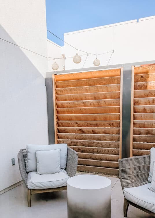 Two cushioned lounge chairs and a round side table create a private outdoor patio seating area enclosed by modern wood slat privacy screens. Decorative globe string lights hang overhead against a bright blue sky, giving the contemporary space a quiet and inviting feel.
