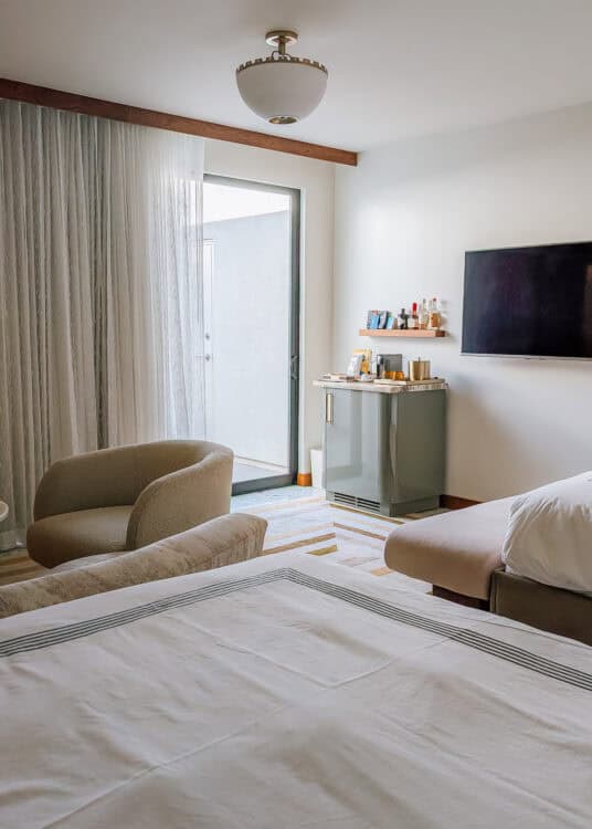 A modern hotel room features two beds with crisp white linens, a curved lounge chair, wall mounted television, and a compact minibar with bottles and coffee supplies. Sheer curtains filter natural light from the patio door, creating a calm and contemporary space designed for relaxation.