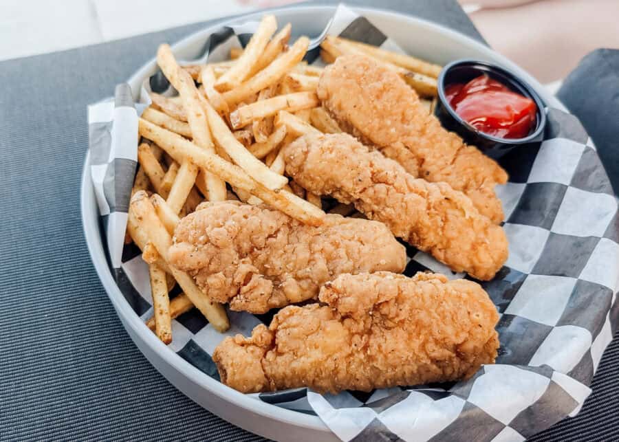 A basket lined with black and white checkered paper holds four crispy fried chicken tenders and a generous serving of seasoned French fries with a side of ketchup for dipping. The golden brown meal is served in a casual dining setting and fills the basket from edge to edge.