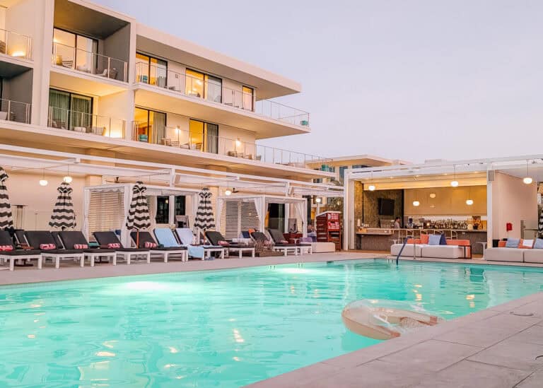 Illuminated pool at Thompson Palm Springs surrounded by modern lounge chairs, striped umbrellas, private cabanas, and an open air bar at dusk. Warm lights from the hotel balconies reflect across the turquoise water, creating a stylish desert resort atmosphere.