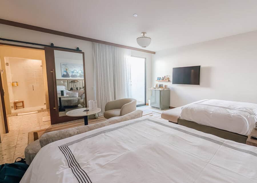 A bright contemporary hotel room includes two queen beds with white linens, a cozy seating area with a round table and upholstered chair, and a wall mounted television beside a stocked minibar. An open bathroom with a glass shower, large sliding mirror, and patio door adds to the spacious, modern design.