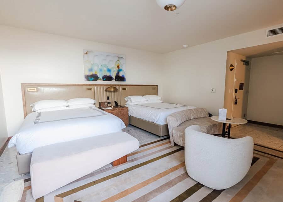 A spacious hotel room features two neatly made queen beds with upholstered headboards, a shared nightstand with brass lighting, and a soft neutral color palette. A curved lounge chair, matching bench, abstract artwork, and striped area rug create a modern, upscale atmosphere with plenty of open space for relaxing.