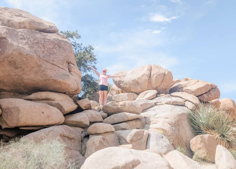 A woman in a pink tank top, black shorts, and an orange cap stands with her arms outstretched on massive granite boulders in Joshua Tree National Park beneath a bright blue sky. The towering rock formations and desert vegetation showcase one of the park's most popular landscapes for hiking and exploring.