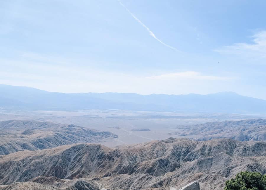 Panoramic view from Mount San Jacinto overlooking the desert valleys and rugged mountain ranges surrounding Palm Springs beneath a clear blue sky with wispy clouds. The sweeping landscape showcases the dramatic contrast between the high alpine peaks and the Coachella Valley below.