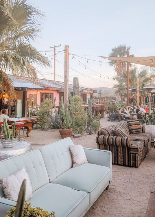 Outdoor courtyard at a desert restaurant with vintage sofas, potted cacti, string lights, and rustic buildings creating a relaxed gathering space at sunset. Palm trees, mountain views, and Southwestern decor add to the inviting atmosphere for dining and drinks in the California desert.