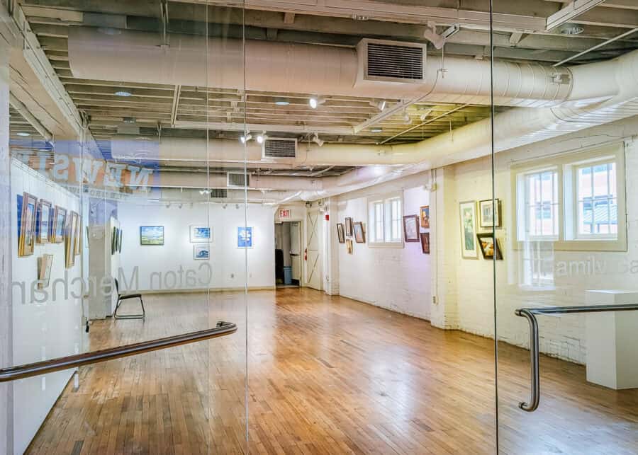 A bright art gallery with polished hardwood floors, exposed ceiling beams and ductwork, and white brick walls displays framed paintings under track lighting. The view through the glass entrance doors shows an open exhibition space with large windows letting in natural light and the words "Next Step" and "Family Gallery" visible on the glass.