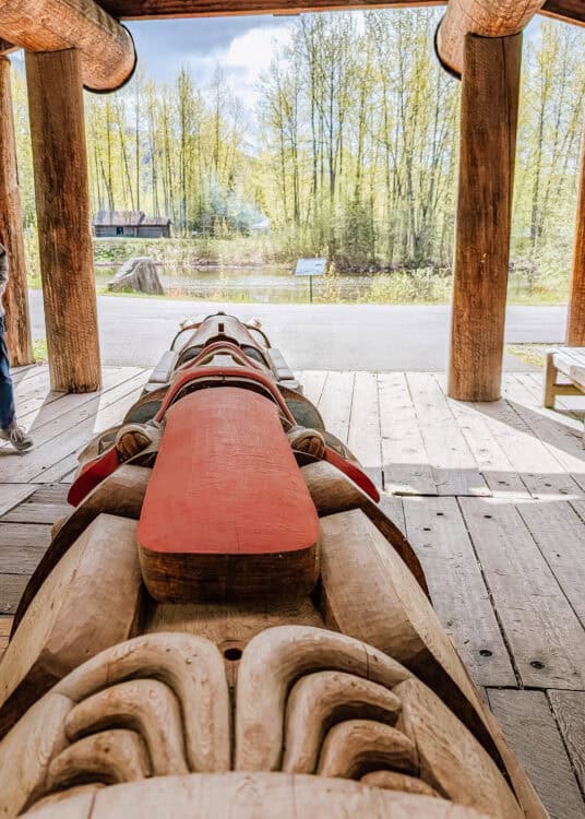 A long carved wooden totem pole with red painted details lies horizontally beneath a rustic timber shelter. Intricate carved patterns fill the foreground with a pond and leafy forest visible beyond.