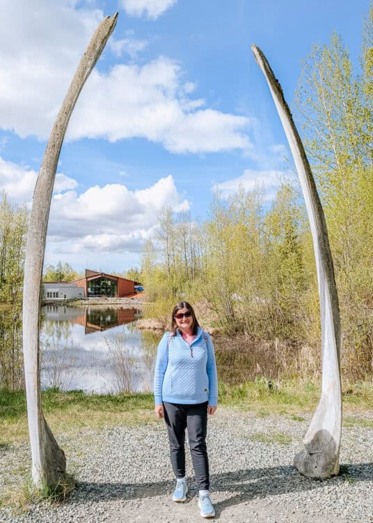 A woman stands between two towering whale bones beside a pond surrounded by spring greenery in Anchorage Alaska. The enormous curved bones frame her against a bright blue sky and show their impressive scale.