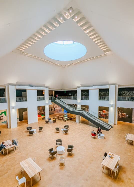 Spacious atrium inside a museum in Anchorage with a circular skylight centered in the high white ceiling. An escalator crosses the open gallery above tables and seating areas with exhibits lining the surrounding walls.
