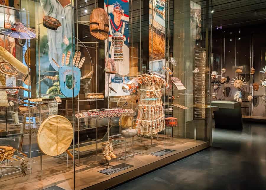 Museum display in Anchorage featuring Alaska Native clothing masks tools and ceremonial objects arranged inside a large glass case. A fur garment decorated with red accents stands near a large round drum while carved masks and other cultural objects fill the surrounding exhibits.