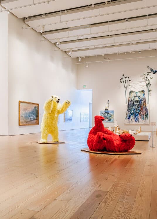 Contemporary art gallery in an Anchorage museum featuring shaggy polar bear sculptures including one white bear standing upright and a bright red polar bear reclining on a low platform. Paintings on the surrounding walls include a large colorful bear portrait.
