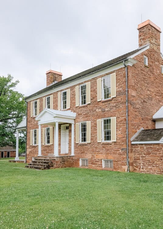 A two story historic stone house with cream shutters, tall brick chimneys, and a white columned front porch sits on a wide grassy lawn beneath an overcast sky. The symmetrical architecture and well preserved masonry highlight the home's historic character and traditional design.
