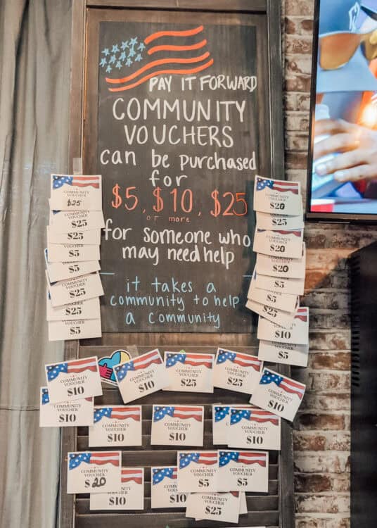 A chalkboard display invites customers to support a "Pay It Forward" community voucher program, with donation cards attached around the sign in amounts of $5, $10, $20, and $25. The sign reads, "PAY IT FORWARD. COMMUNITY VOUCHERS can be purchased for $5, $10, $25 or more for someone who may need help. It takes a community to help a community," highlighting a restaurant initiative to provide meals or assistance for others.